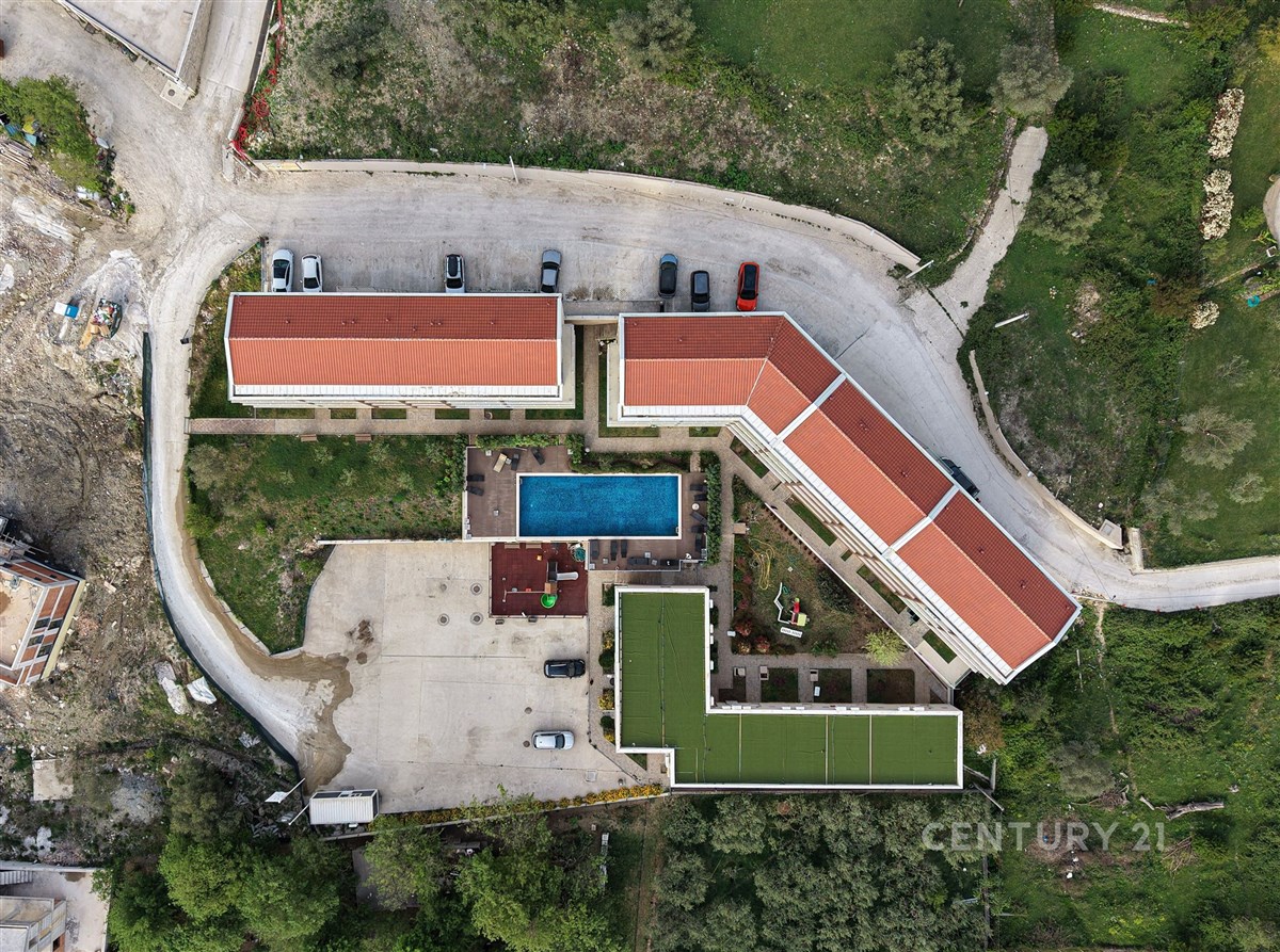 Letecký pohled na moderní bytový komplex s červenou střechou a venkovním bazénem, obklopený zelení, parkovištěm a svěžími zahradami.
