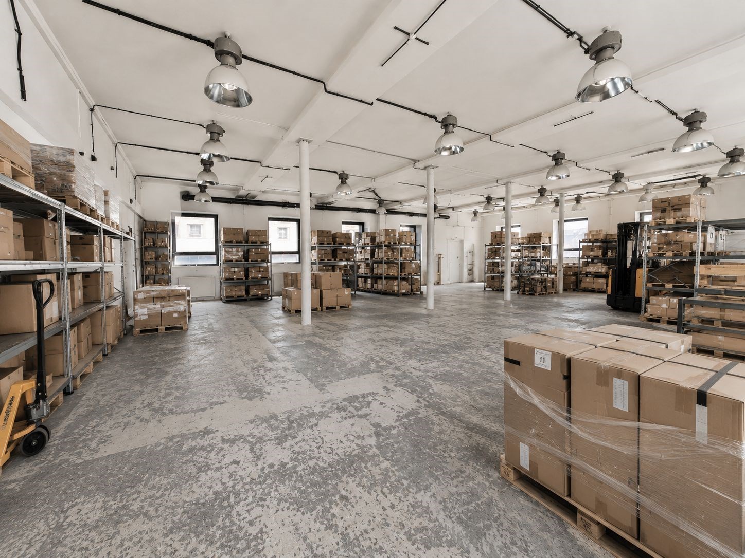 Skladový prostor s betonovou podlahou, vysokým stropem a přirozeným osvětlením, vybaven regály pro skladování, ideální pro logistiku nebo výrobu.