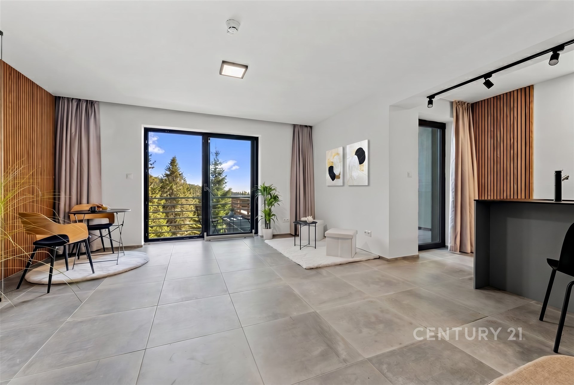 Světlý obývací pokoj s velkými okny, dlažbou a moderním designem, propojený s kuchyňským koutem, nabízí výhled na přírodu.