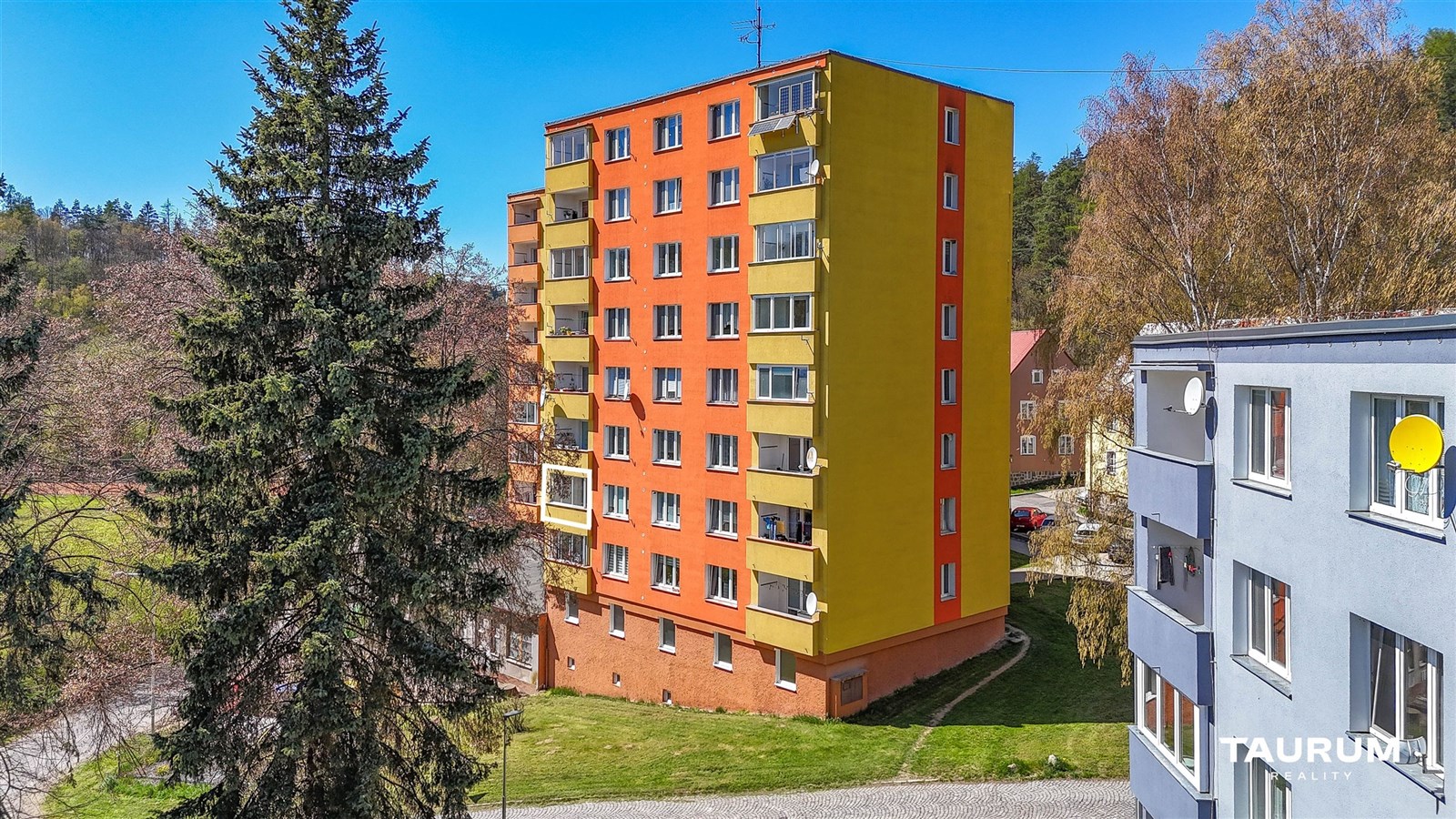 Barevná fasáda panelového domu s balkony obklopena zelení, zrekonstruovaná budova ve světlém stylu, klidná lokalita poblíž lesa.
