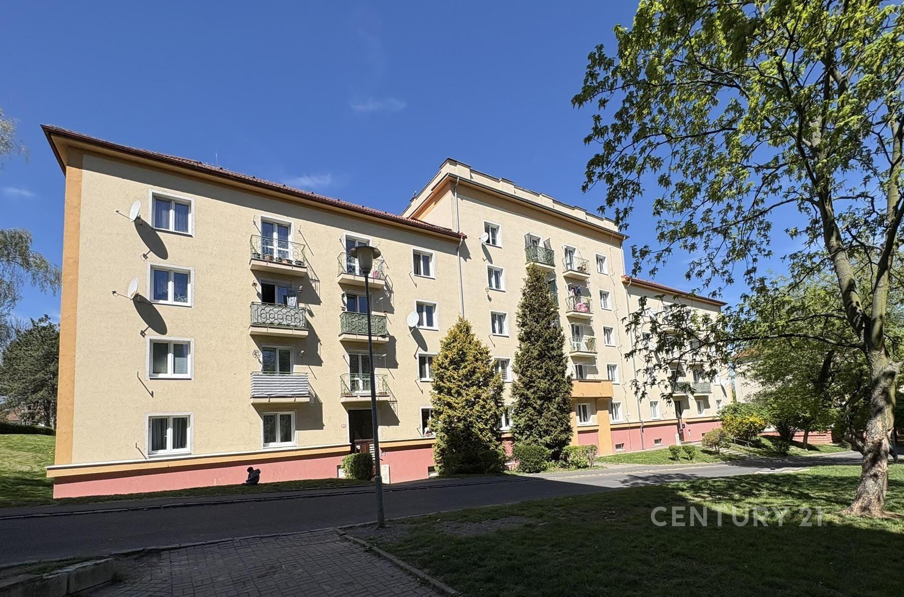Čtyřpatrový bytový dům s balkony, okna s bílými rámy, obklopen zelení, v klidné rezidenční čtvrti, dobrý stav, cihlová fasáda.