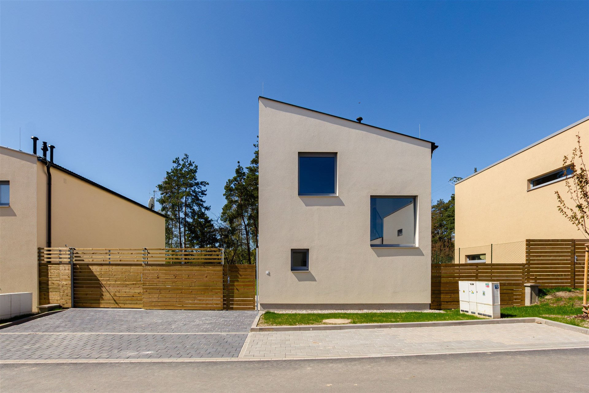 Moderní rodinný dům s minimalistickou fasádou, velkými okny a dřevěným plotem, nachází se v klidné vilové čtvrti.