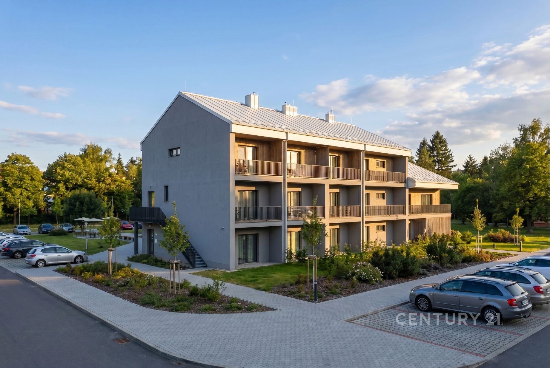 Moderní bytový dům s velkými okny a dřevěnými balkony, zasazený v klidné rezidenční čtvrti s asfaltovým parkovištěm.