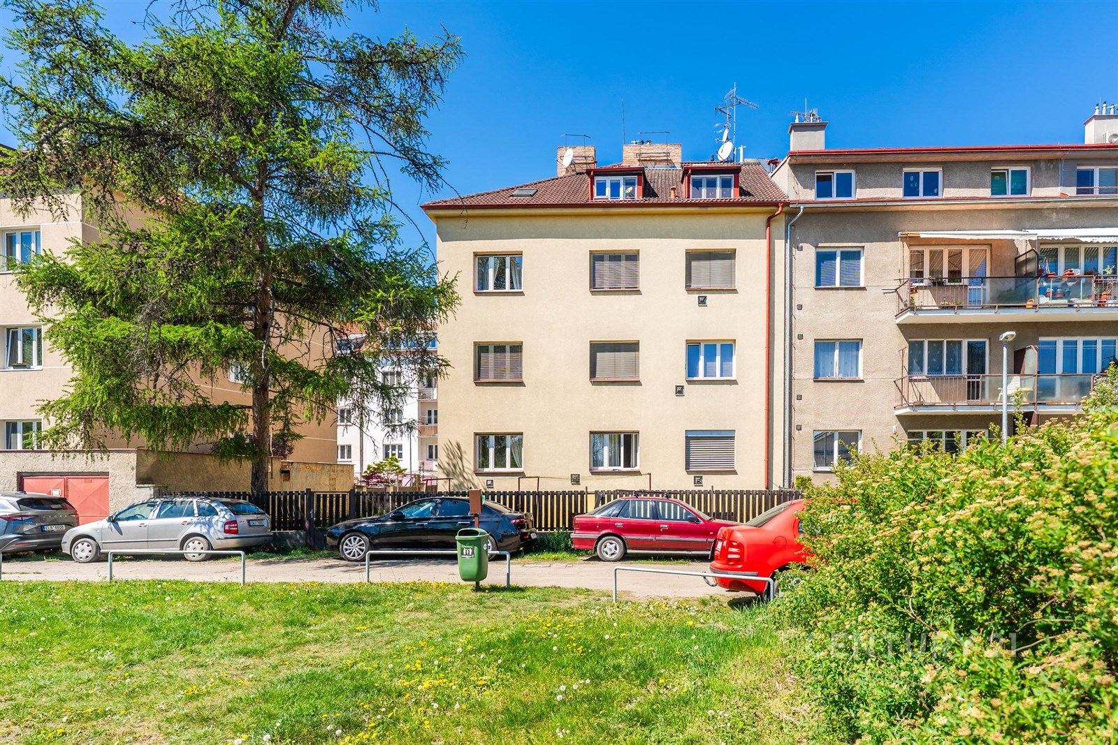 Exteriér bytového domu s udržovanou zahradou, parkováním a červeným autem; ideální lokalita s klidným okolím, vhodná pro rodiny.