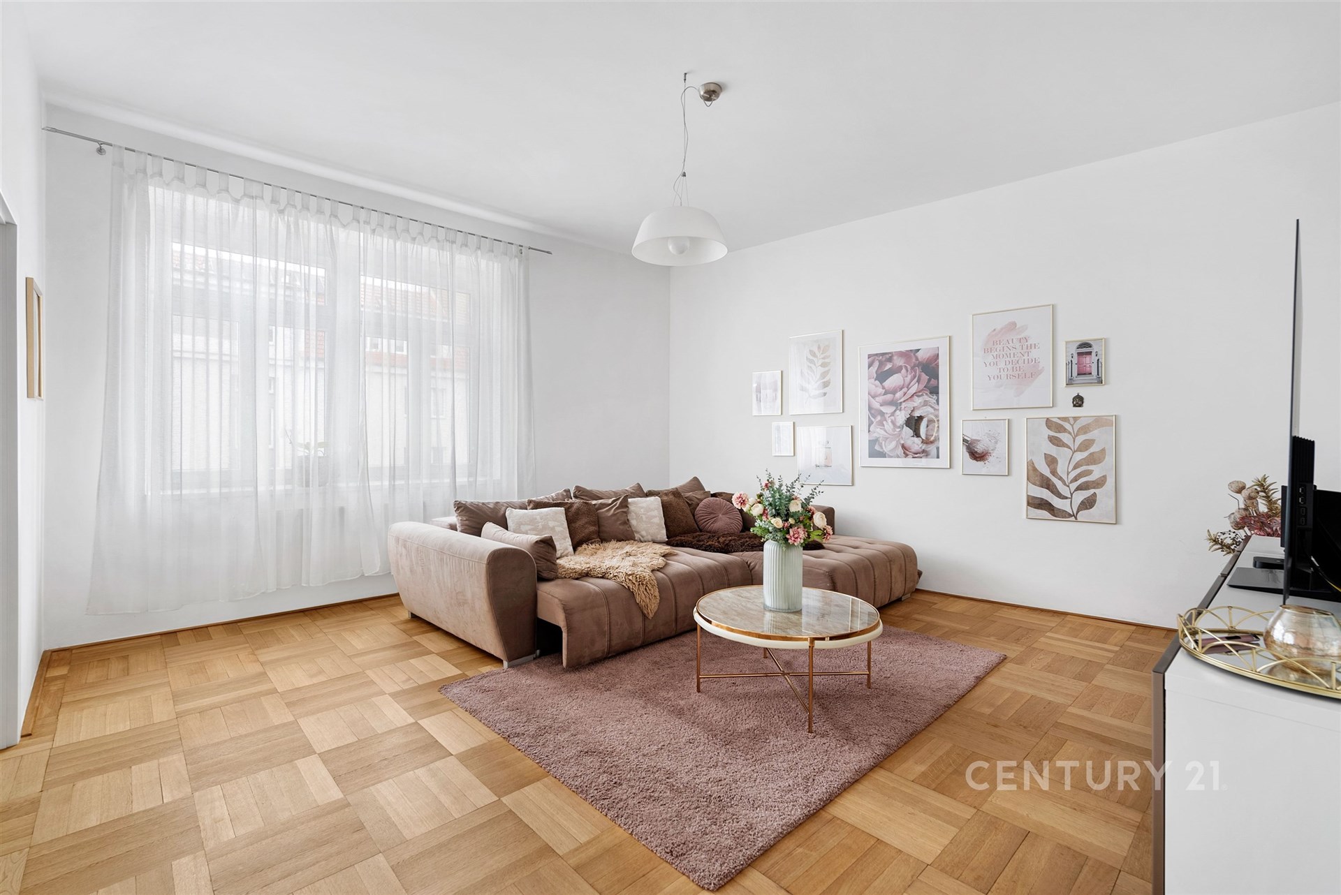 Světlý obývací pokoj s parketovou podlahou, velká okna a moderní výzdoba, která vytváří útulný a elegantní prostor.