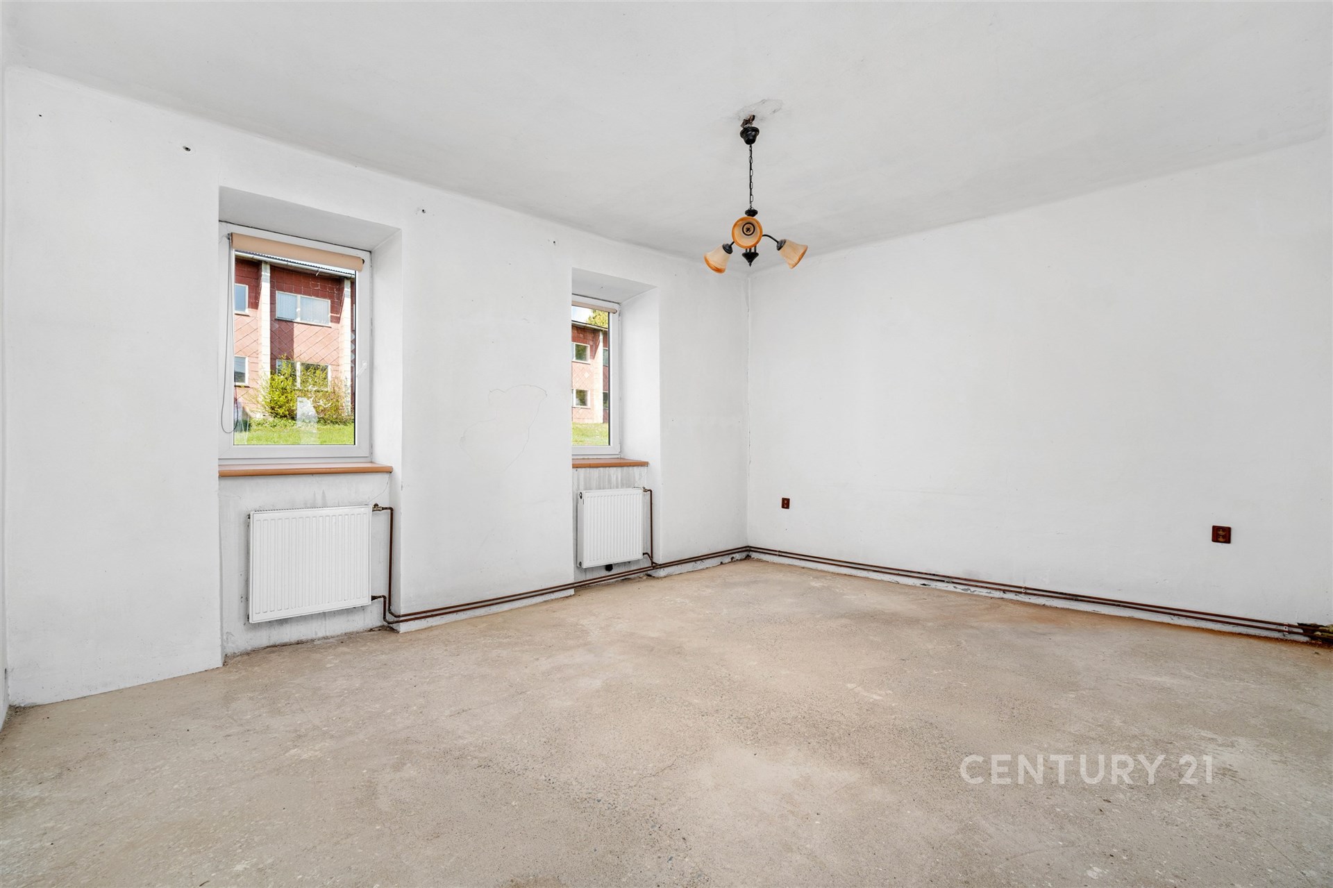 Prázdná světlá místnost, betonová podlaha, bílá výmalba, dvě okna s výhledem na zeleň, příprava na rekonstrukci, ideální pro modernizaci