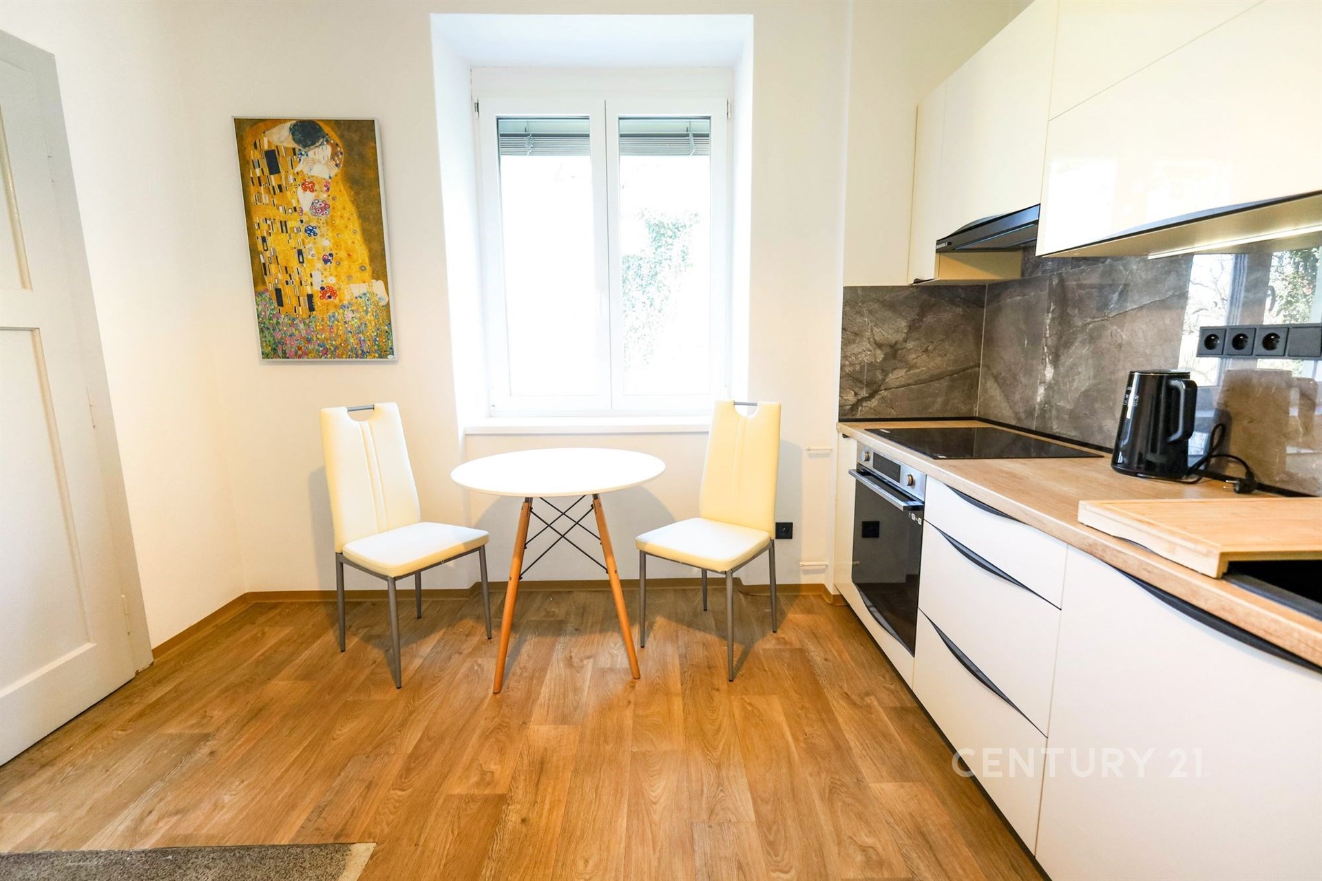 Kuchyň s moderním nábytkem, parketová podlaha, světlý prostor, kachlová zástěna a velké okno poskytují přirozené světlo