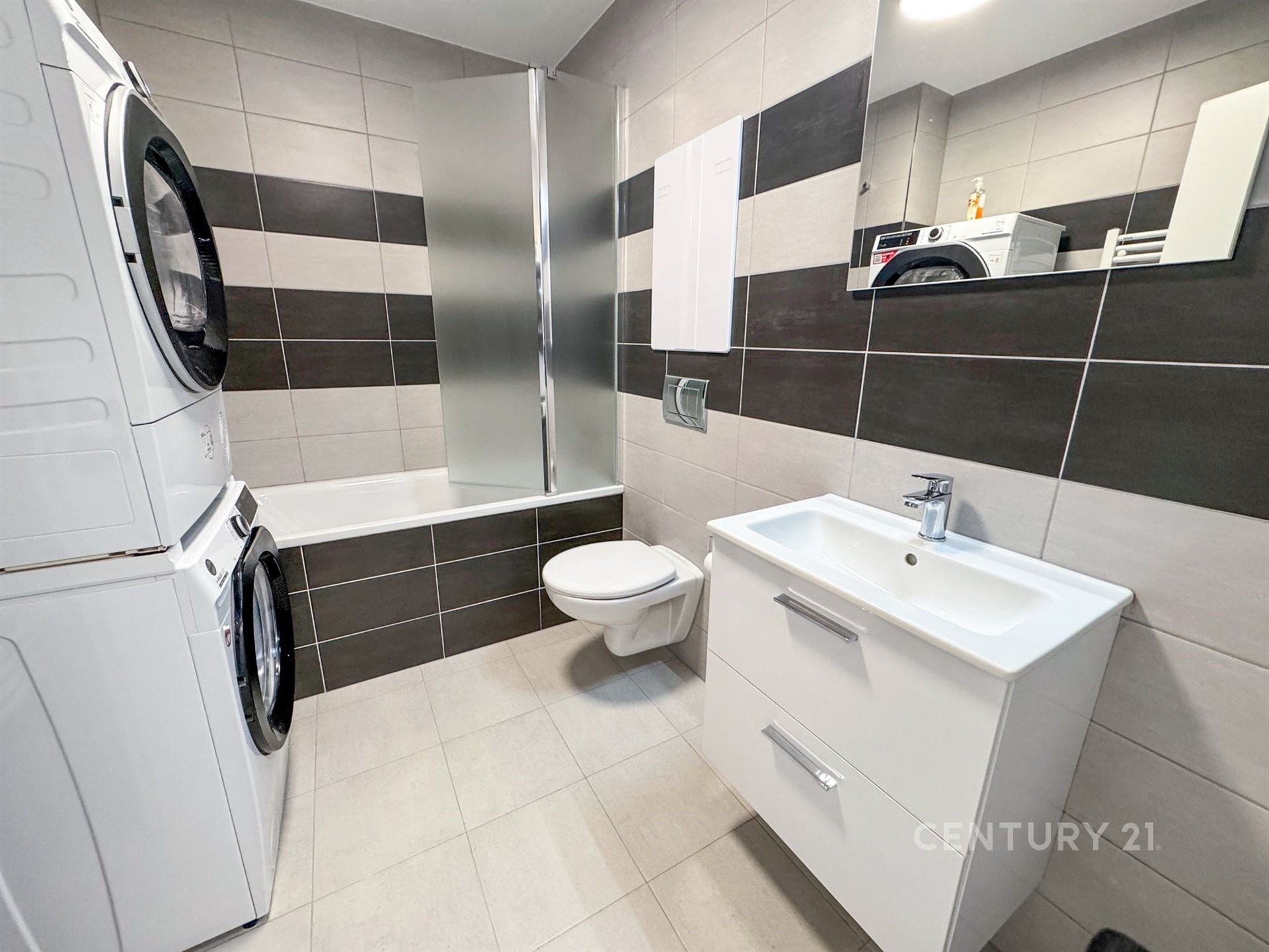 Moderní koupelna s dlažbou, vestavěným umyvadlem a úložnou skříňkou. Kombinace světlých a tmavých obkladů, pračka se sušičkou.