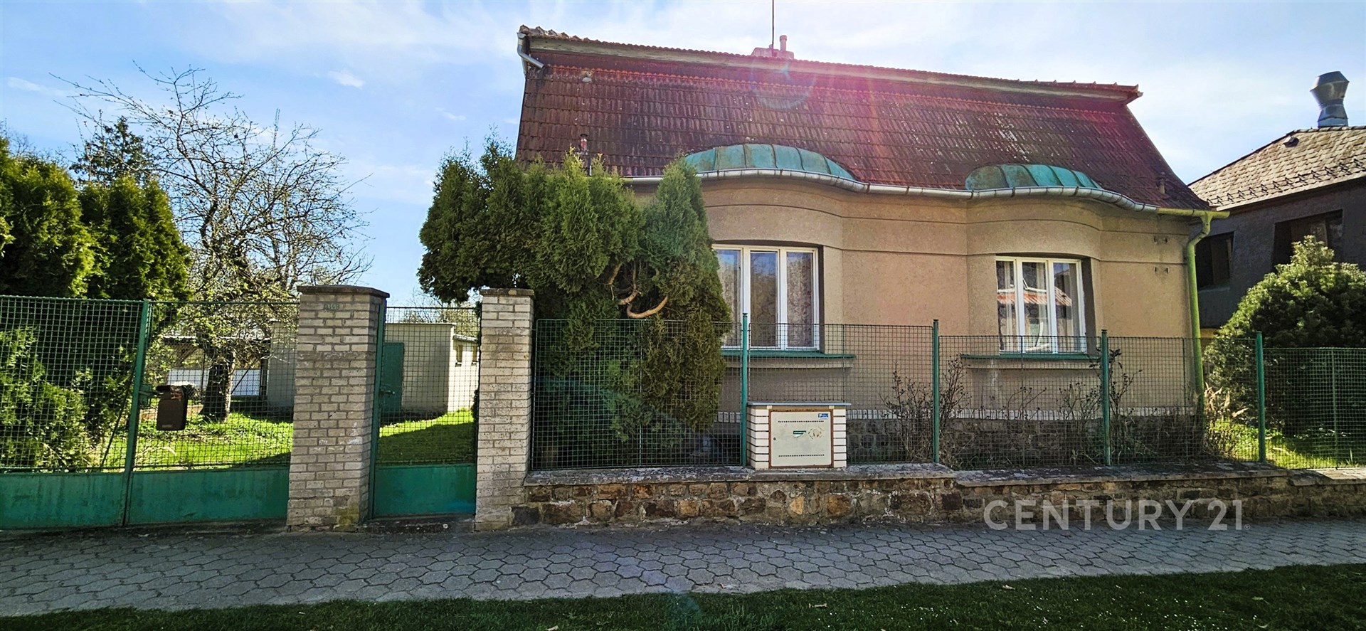 Rodinný dům s šikmou střechou a zeleným plotem, obklopený zahradou. Okna s oblouky, klidná lokalita.