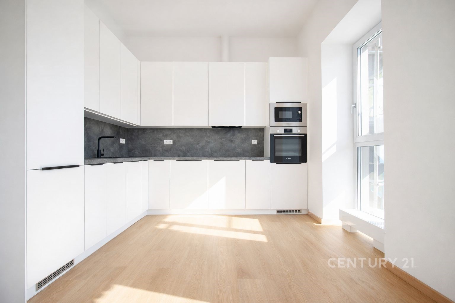Moderní kuchyň s parketovou podlahou, velká okna zajišťující světlý interiér, bílá kuchyňská linka s vestavěnými spotřebiči.