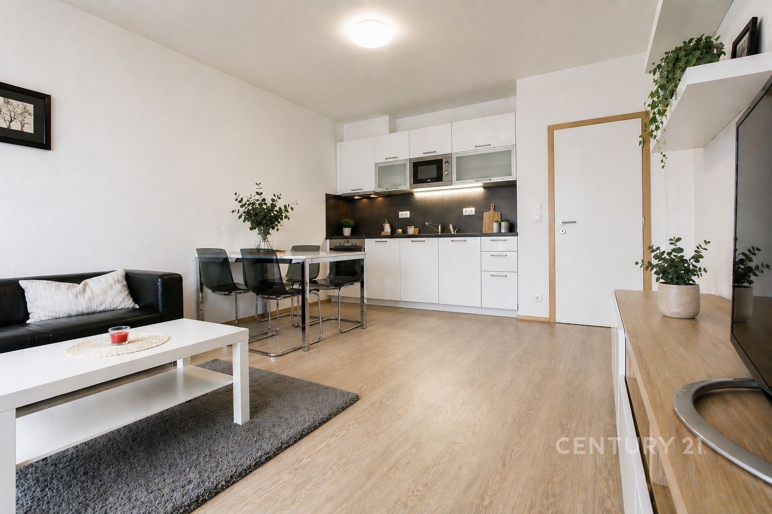 Světlý obývací pokoj s dřevěnou podlahou, moderní kuchyňskou linkou a velkými okny, včetně vestavěného úložného prostoru a elegantního nábytku.