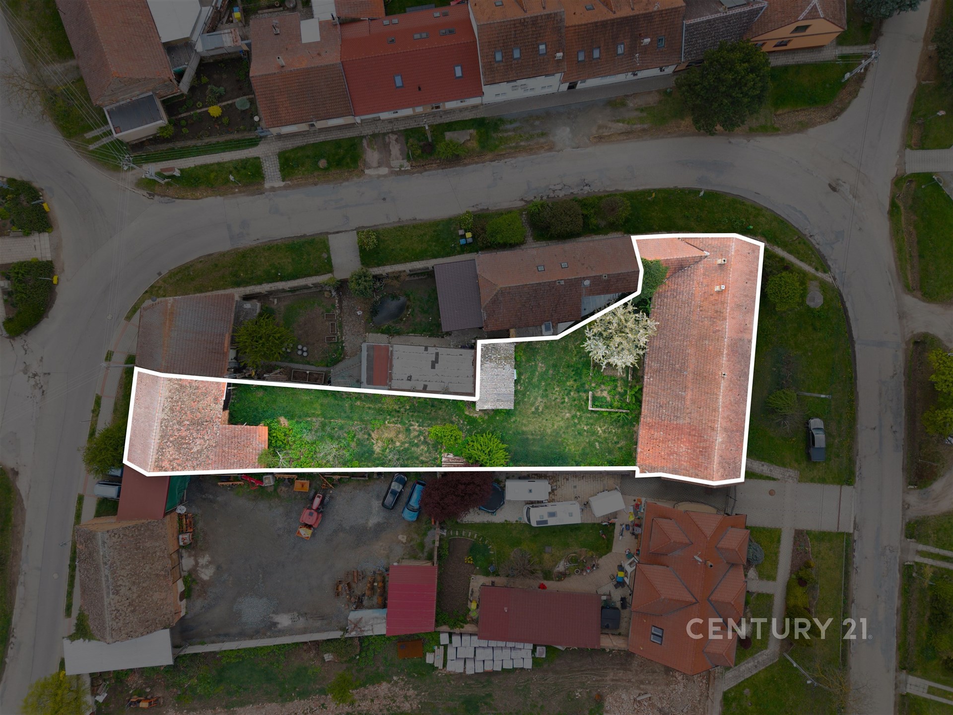 Pohled z ptačí perspektivy na pozemek s dvěma stavbami a zahradou, umístěný v klidné rezidenční oblasti s červenými střechami domů.