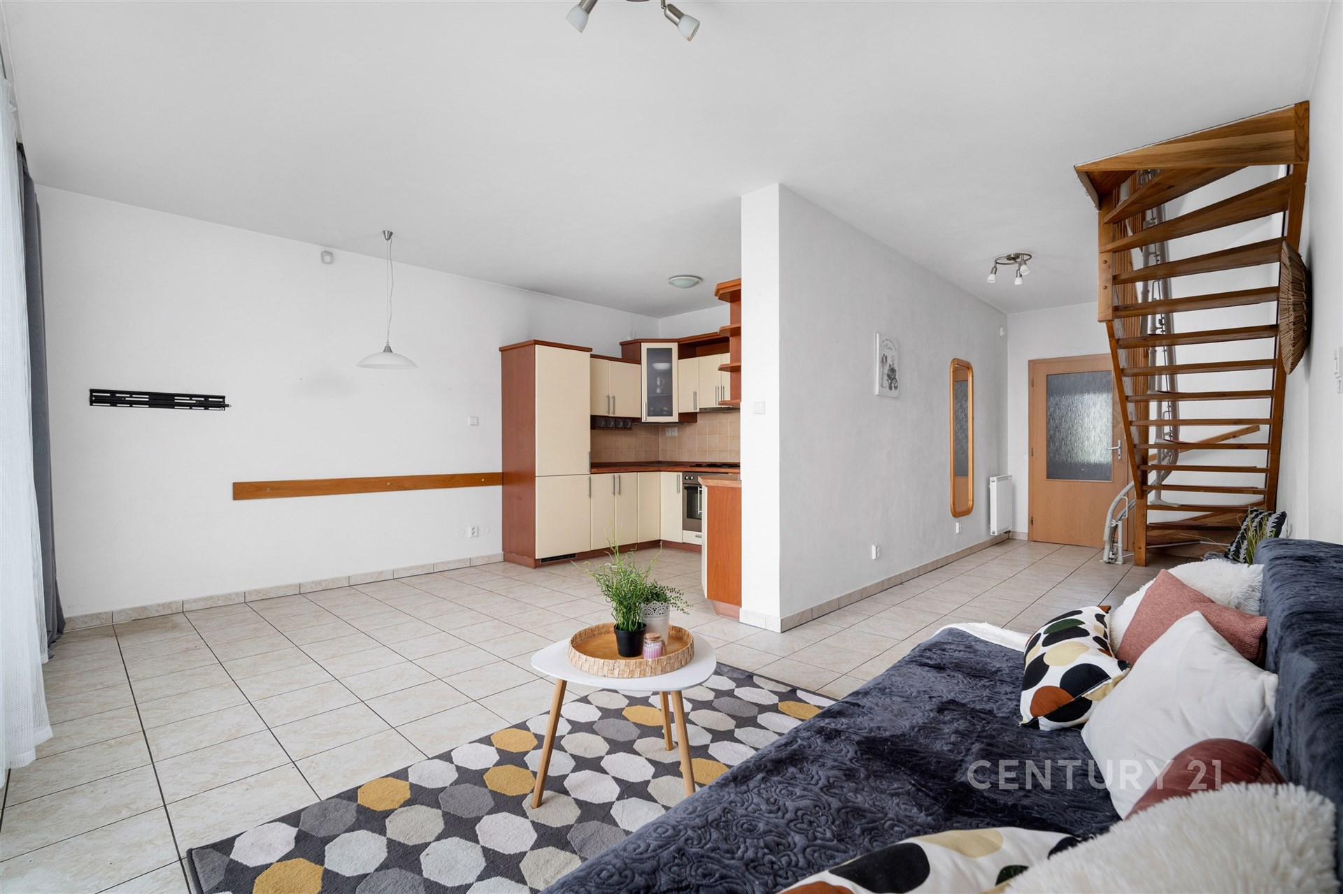 Obývací pokoj s moderní dlažbou, dřevěným schodištěm a světlou kuchyní. Velká okna umožňují přirozené světlo a vytváří prostorný dojem.