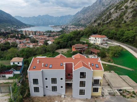 Moderní bytový dům s červenou střechou a velkými okny, nacházející se v malebné oblasti s výhledem na hory a nedaleké jezero.