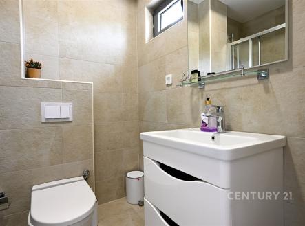 Koupelna s moderními dlaždicemi, světlé barvy, závěsné WC, velké zrcadlo, praktická skříňka pod umyvadlem.