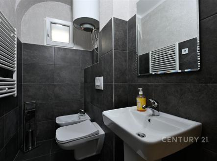 Malá koupelna s moderní dlažbou, závěsné WC a bidet, umyvadlo se zrcadlem, elektrický žebřík a světlé okno propouštějící denní světlo.