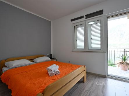 Ložnice s moderním stylem a dřevěnou podlahou, velká okna s výhledem na terasu, dveře vedou na balkon, světlá a vzdušná atmosféra.