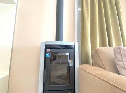 Obývací pokoj s moderními krbovými kamny, světlé stěny a velké okno s dlouhými závěsy, dlažbová podlaha.