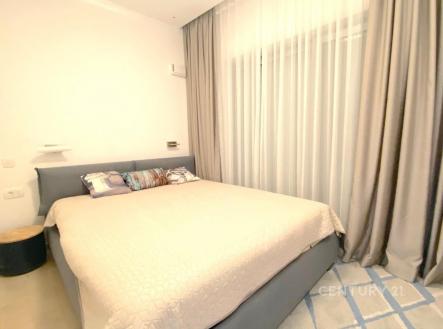 Prostorná ložnice s moderním nábytkem, velká manželská postel, světlé závěsy a parketová podlaha, elegantní a světlý interiér.