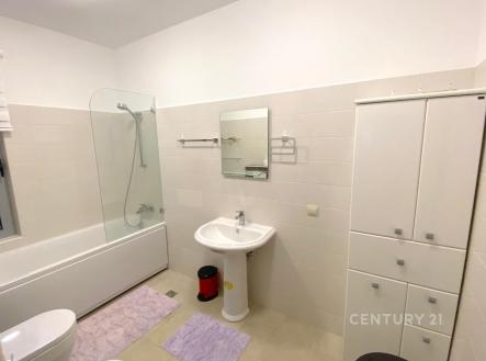 Prostorná koupelna s moderními dlaždicemi, světlou vanou a zrcadlem nad umyvadlem, nabízí praktické úložné prostory a elegantní design.