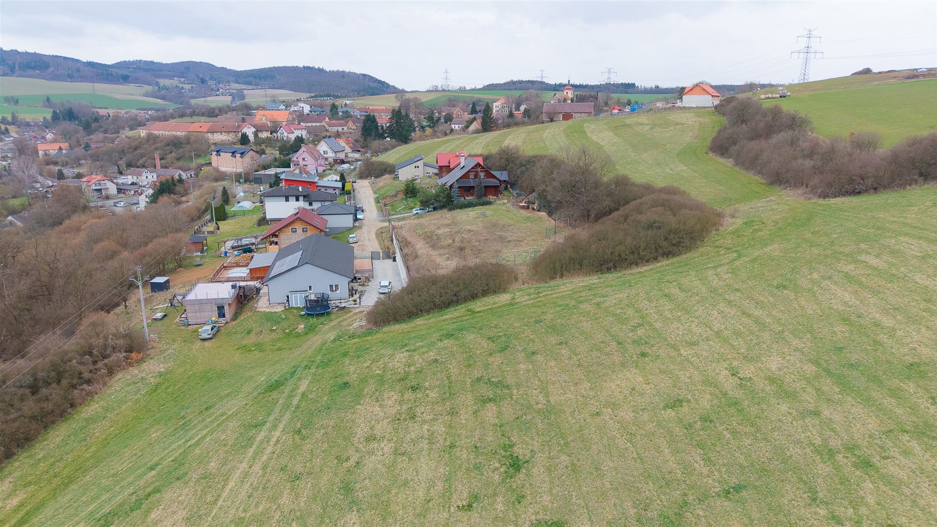 Prázdný stavební pozemek na okraji obce, obklopený zelenými loukami a kopce, ideální pro výstavbu rodinného domu, klidná lokalita
