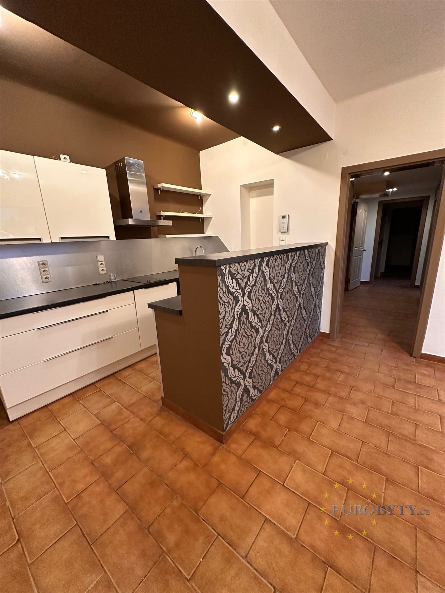 Moderní kuchyň s dlažbou a vestavěnými skříněmi, moderní nerezová digestoř, vedle prostorná chodba s dalším úložným prostorem.