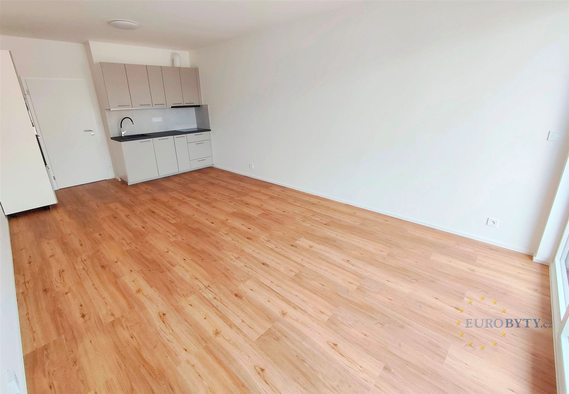 Prostorný obývací pokoj s parketovou podlahou, velká okna umožňující dostatek světla, kuchyňský kout s moderními spotřebiči.