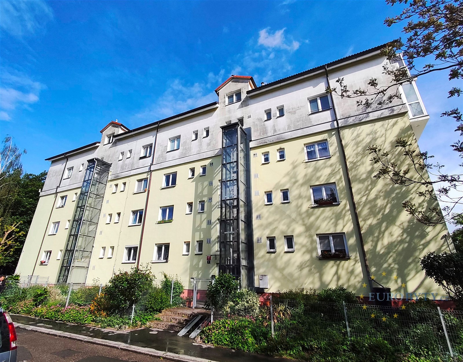 Exteriér bytového domu se světlou fasádou, velká okna a klidná rezidenční lokalita, moderní architektonické prvky, dobrý stav budovy.