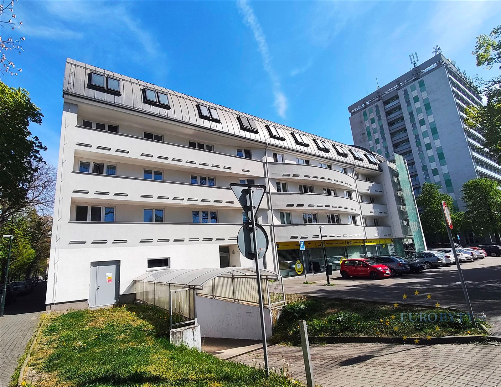 Moderní bytový dům s bílou fasádou a zakřivenou střechou v městském prostředí, nacházející se poblíž hlavní silnice a sousedních budov.