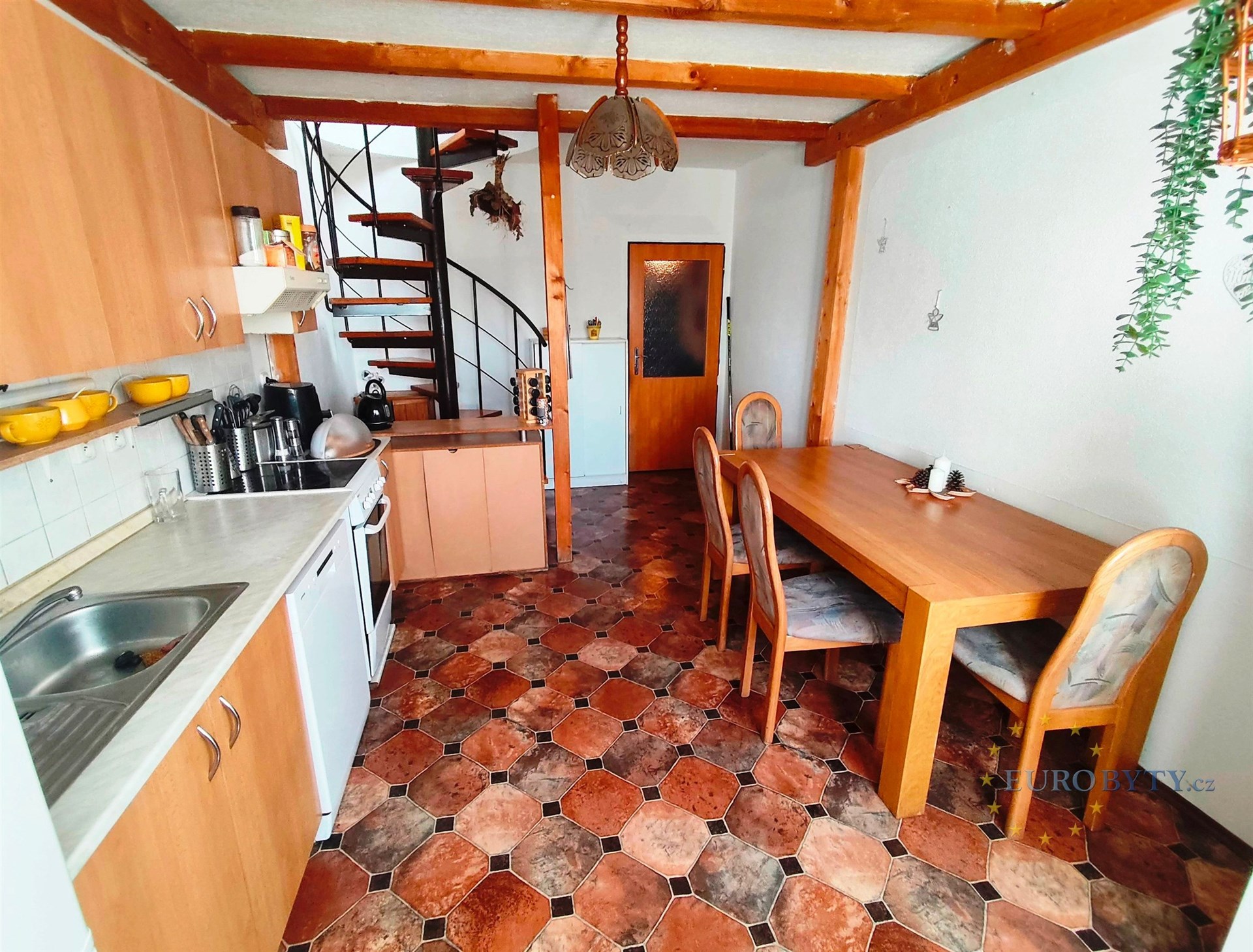 Kuchyň s keramickou dlažbou, dřevěnými stropními trámy a moderní kuchyňskou linkou, vedle rustikálního jídelního stolu se schodištěm.
