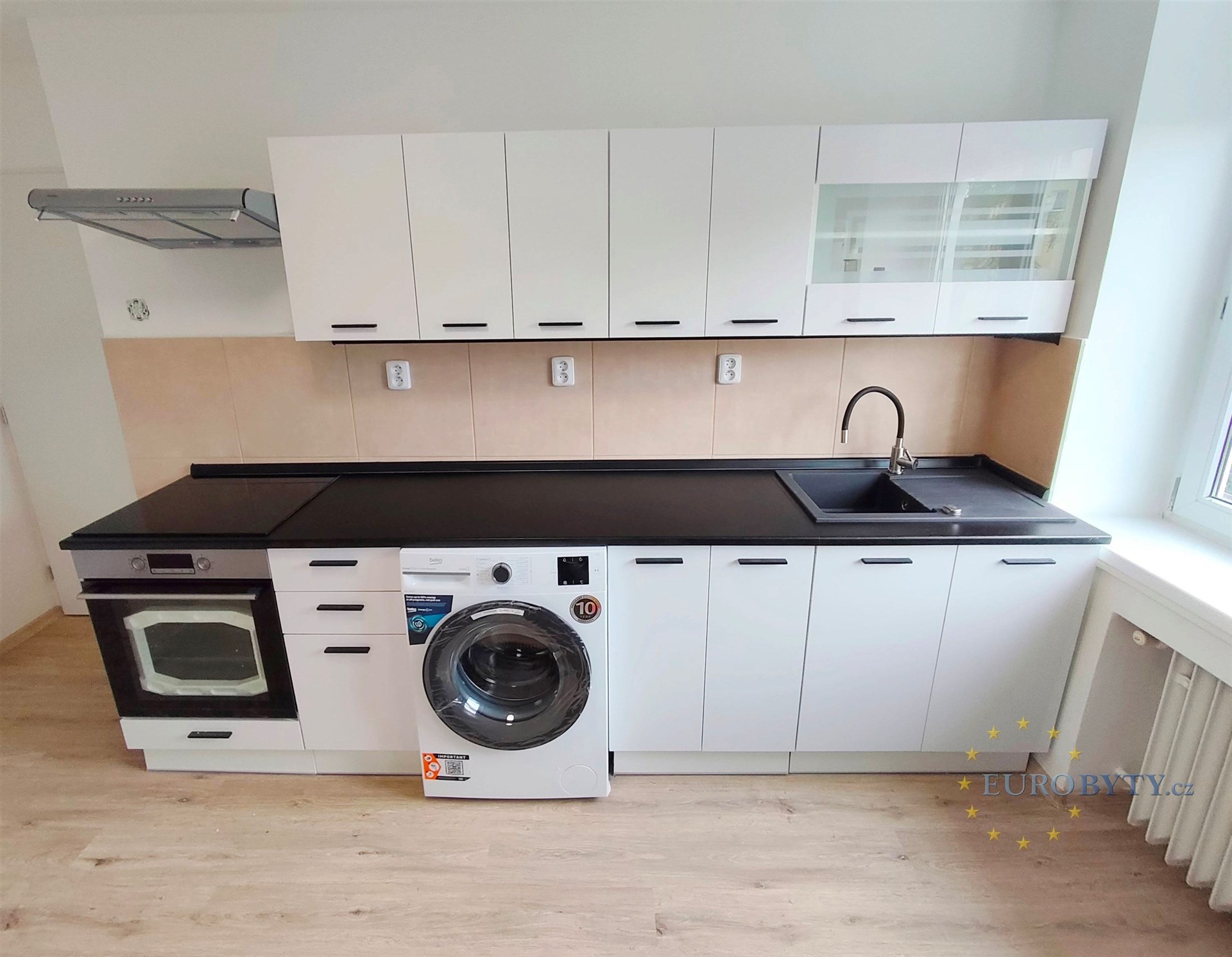 Moderní kuchyň s parketovou podlahou, bílými skříněmi a vestavěnými spotřebiči, velké okno přináší spoustu světla.