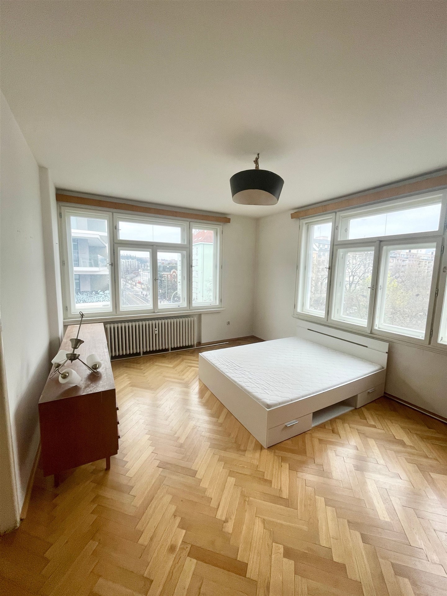 Prostorná ložnice s parketovou podlahou, velká okna, moderní osvětlení, světlý interiér, minimalistický design, vestavěná skříňka.