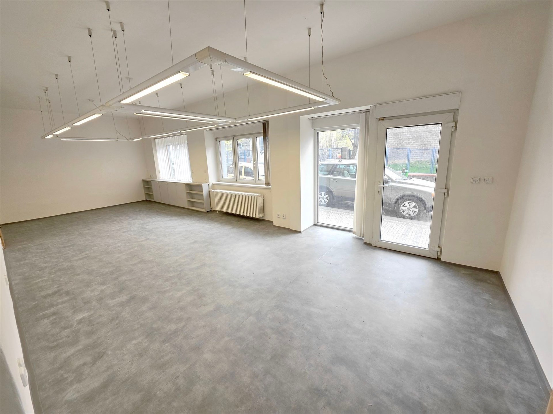 Prázdný kancelářský prostor s betonovou podlahou, velkými okny a moderním osvětlením.