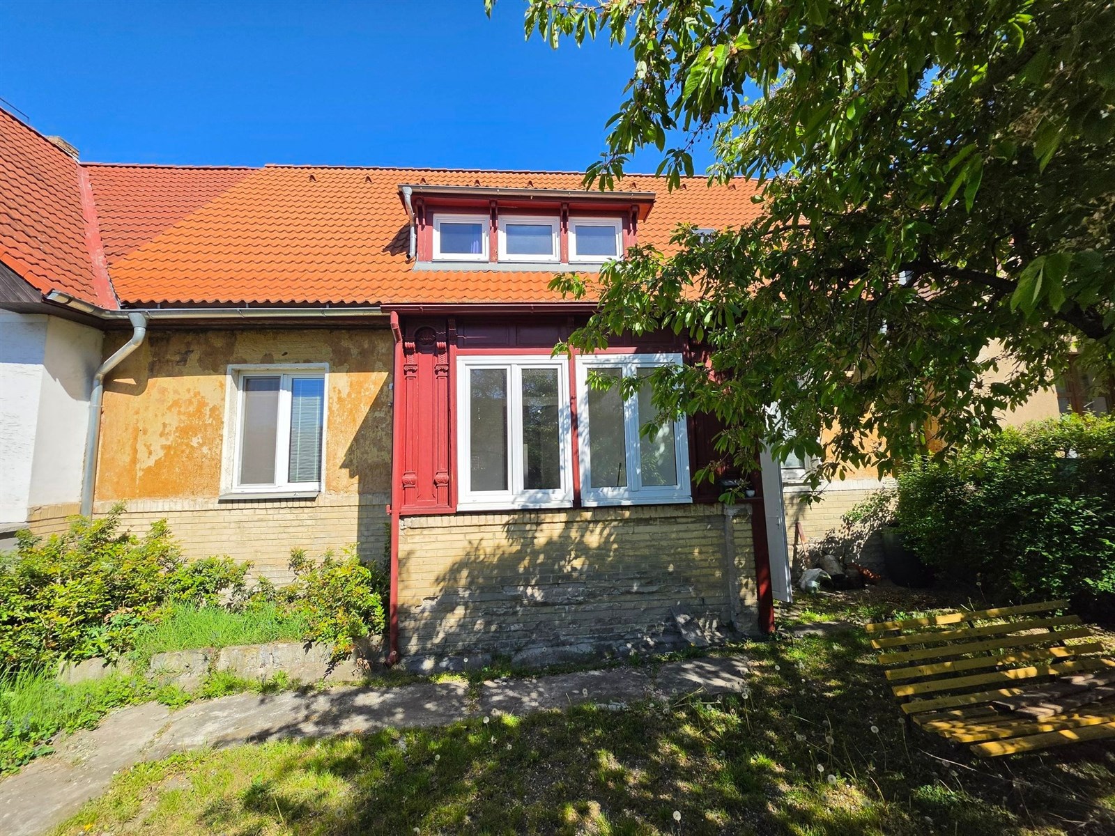 Rodinný dům s červenou střechou, dřevěnými prvky a velkými okny, obklopený zahradou. Nemovitost v klidné lokalitě vhodná pro rekonstrukci.
