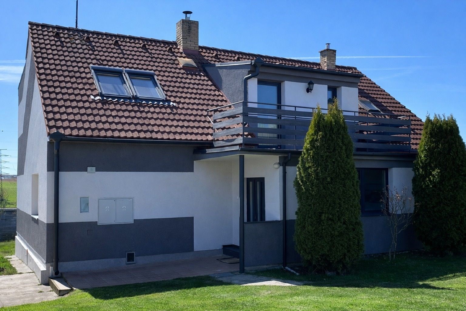 Moderní rodinný dům s červenou střechou, balkónem a zahradou, částečně zateplený, nacházející se na klidném předměstí.