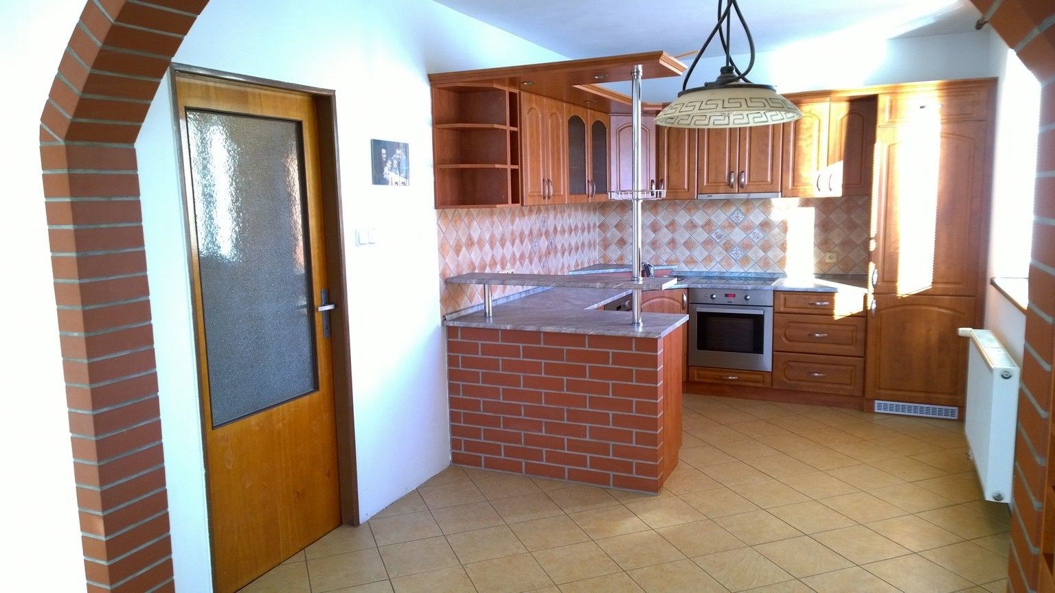 Kuchyň spojená s jídelnou, cihlový pult, dřevěné skříňky, dlažba na podlaze, světlý interiér, rustikální styl.