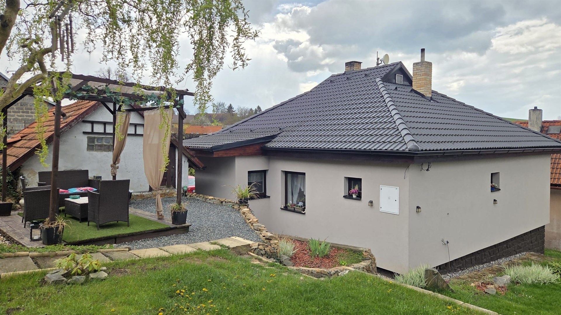 Rodinný dům s moderní fasádou a plechovou střechou, zahrada s terasou a venkovním posezením, klidná lokalita blízko přírody.