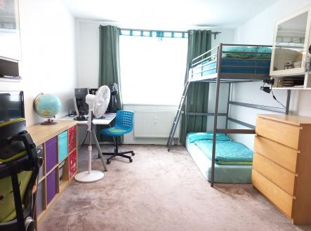 Dětský pokoj s patrovou postelí, kobercovou podlahou a velkým oknem. Moderní interiér nabízí dostatek úložného prostoru pro pohodlné bydlení.