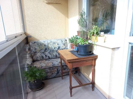 Balkón s dlažbou, pohodlnou sedačkou a dřevěným stolkem. Skleněná zábradlí a rostliny dodávají prostoru útulný a světlý vzhled.