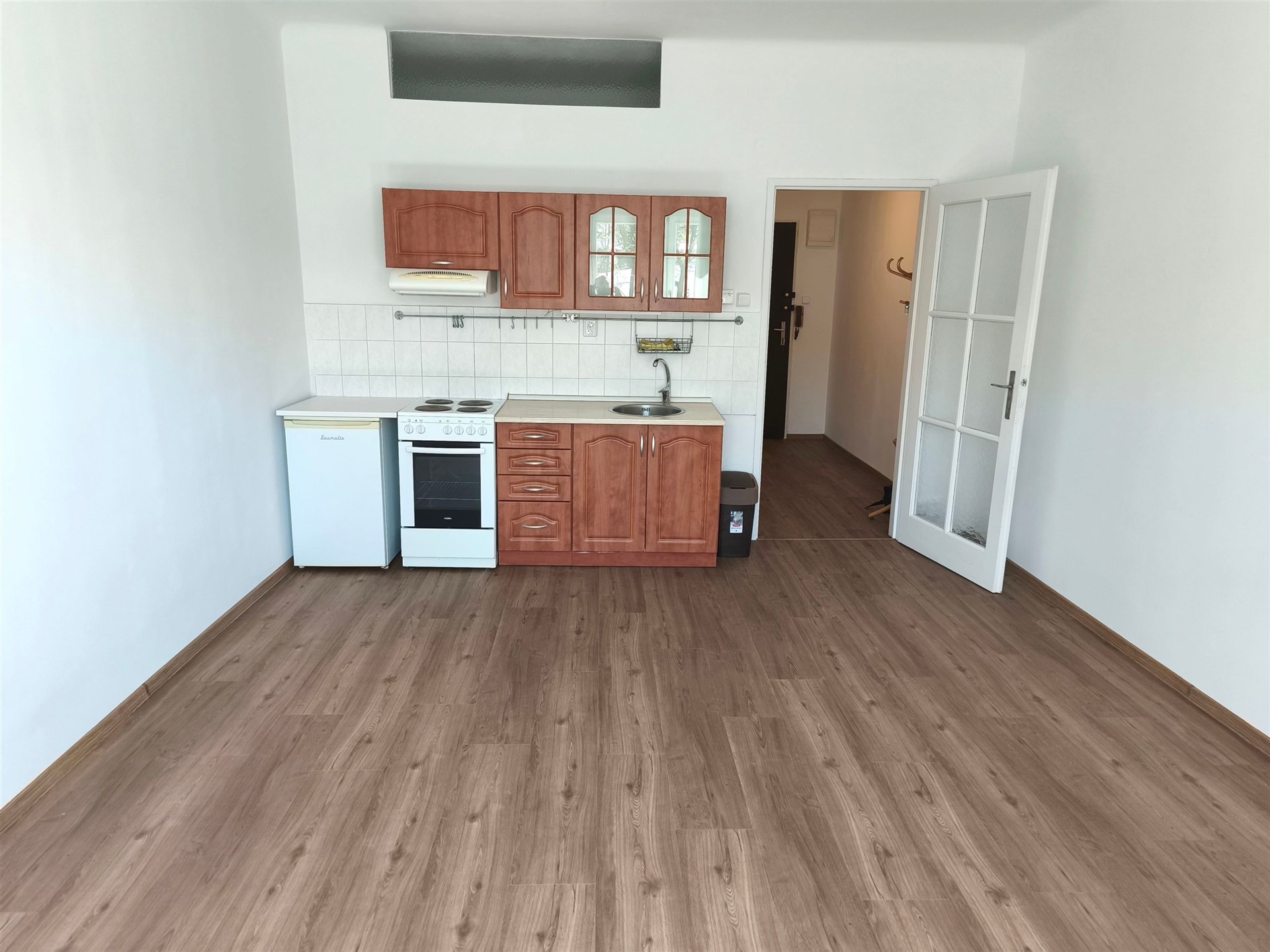 Kuchyňský kout s dřevěnými skříňkami a bílými dlaždicemi, vybavený sporákem a lednicí, na podlaze moderní laminátové parkety.