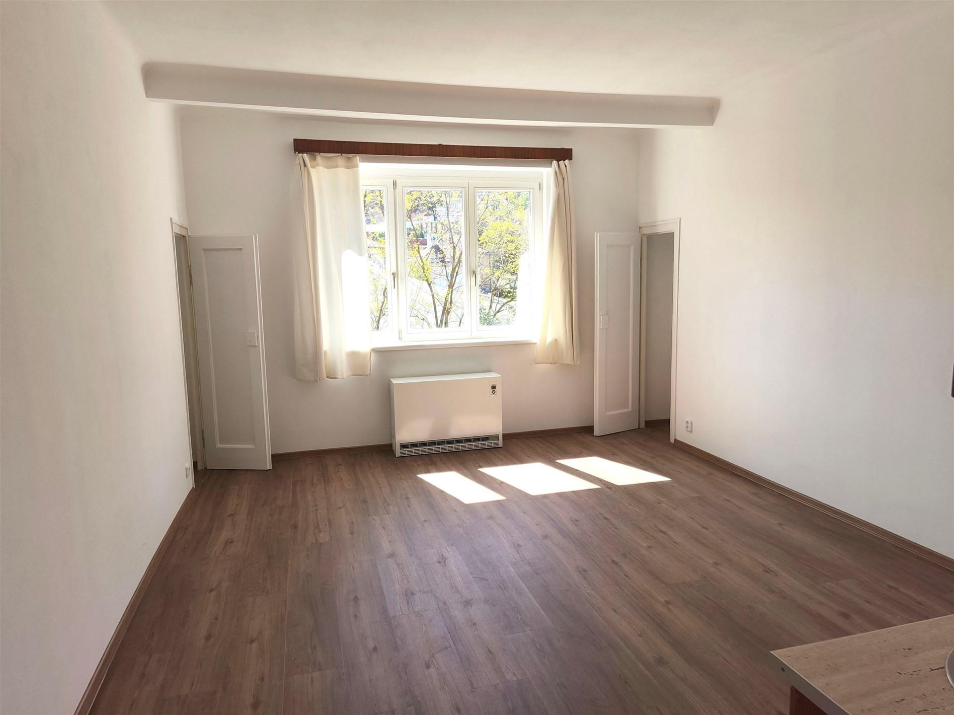 Světlý obývací pokoj s parketovou podlahou, velká okna propouštějí přirozené světlo, moderní prostor ideální pro útulné bydlení.