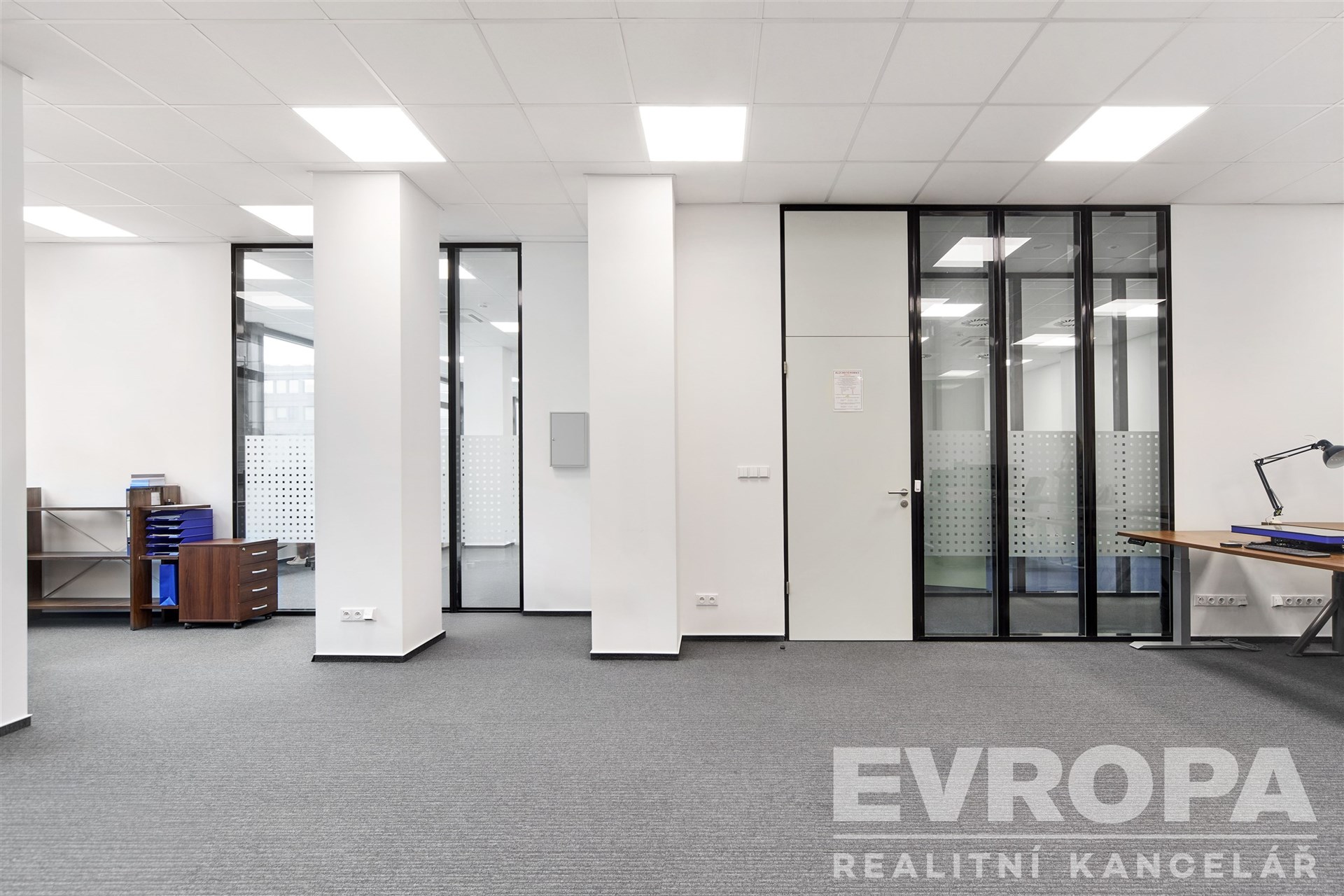 Pracovní kancelář s moderními skleněnými příčkami, šedým kobercem a vestavěným úložným prostorem, ideální pro kreativní týmy.