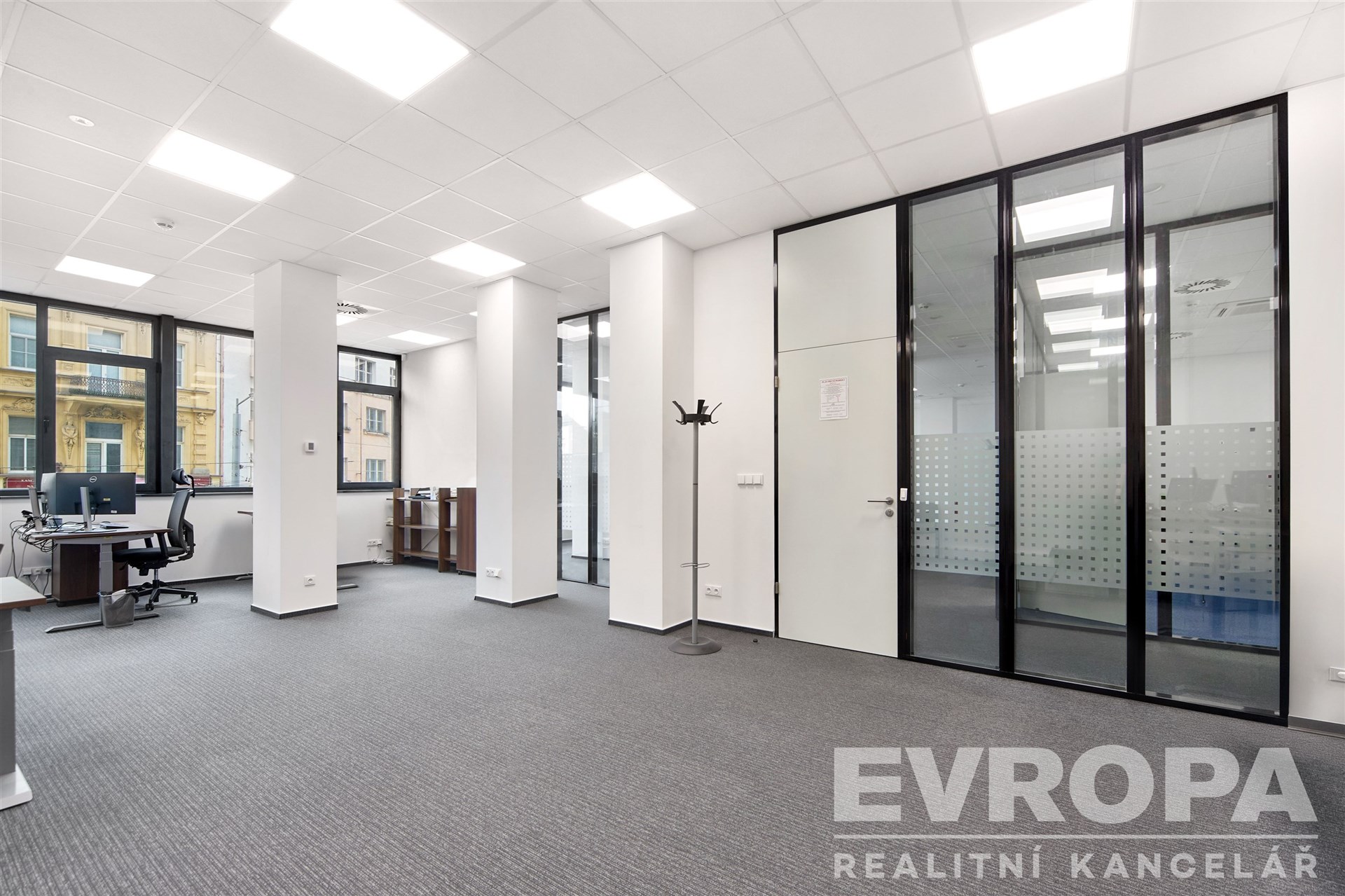 Kancelářský prostor s moderní výzdobou, skleněné příčky, kvalitní koberce, velká okna poskytující dostatek přirozeného světla.