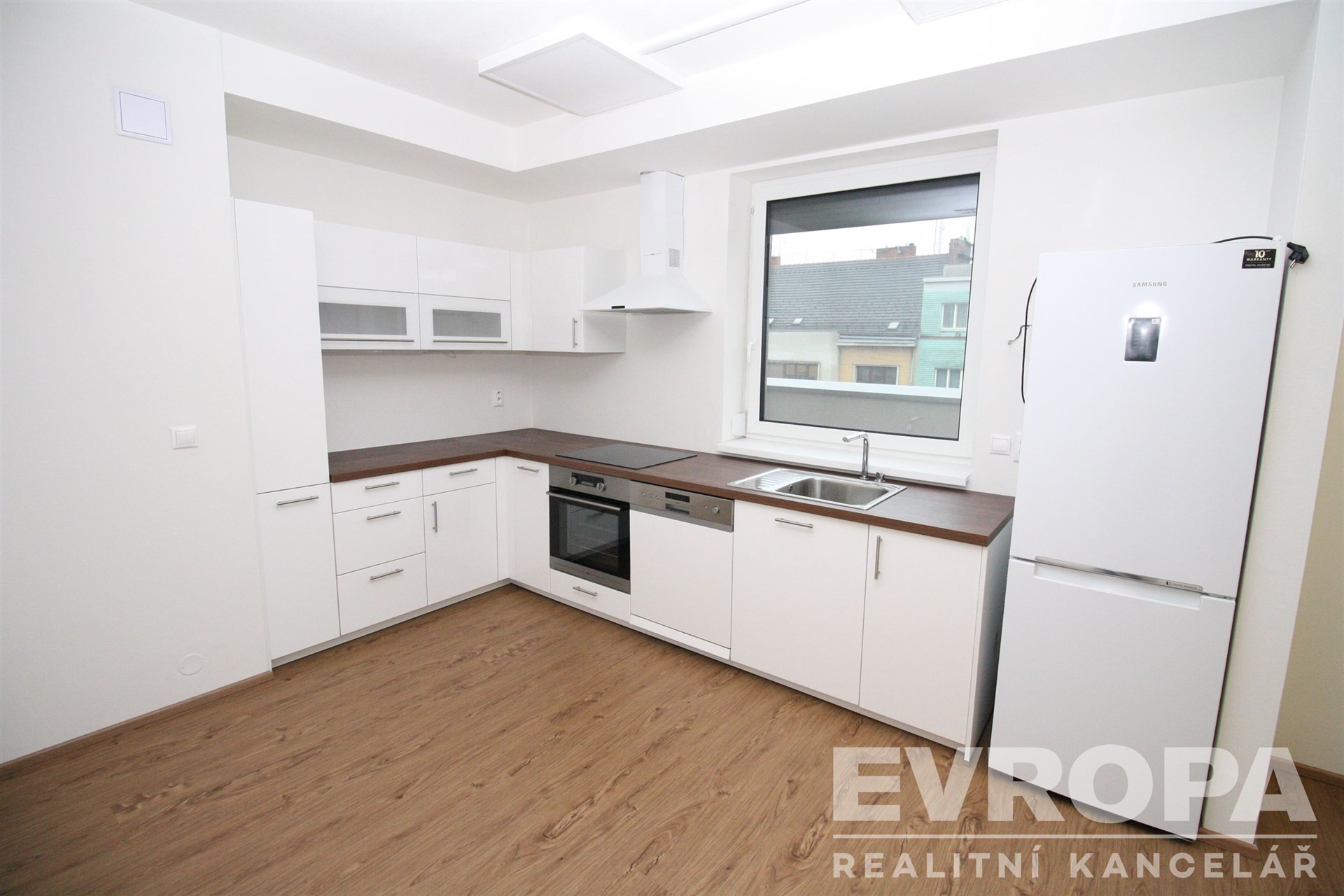 Moderní kuchyň, bílá dvířka, vestavěné spotřebiče, parketová podlaha, velká okna, světlý interiér, elegantní design, kvalitní materiály.