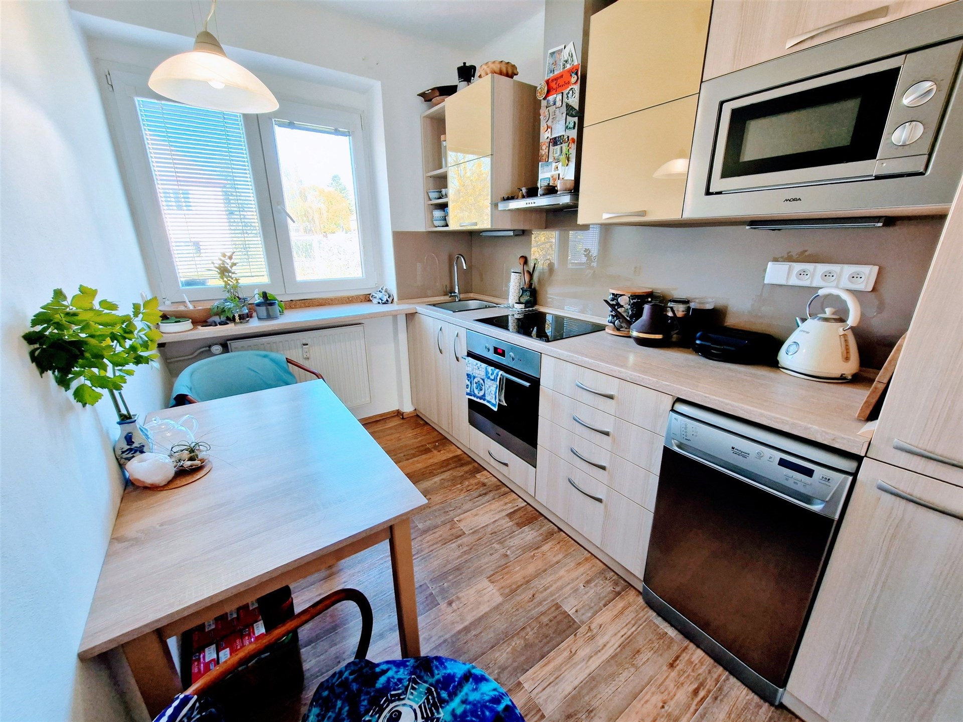 Moderní kuchyň s dřevěnou podlahou, vestavěnými spotřebiči a velkým oknem pro přirozené světlo.