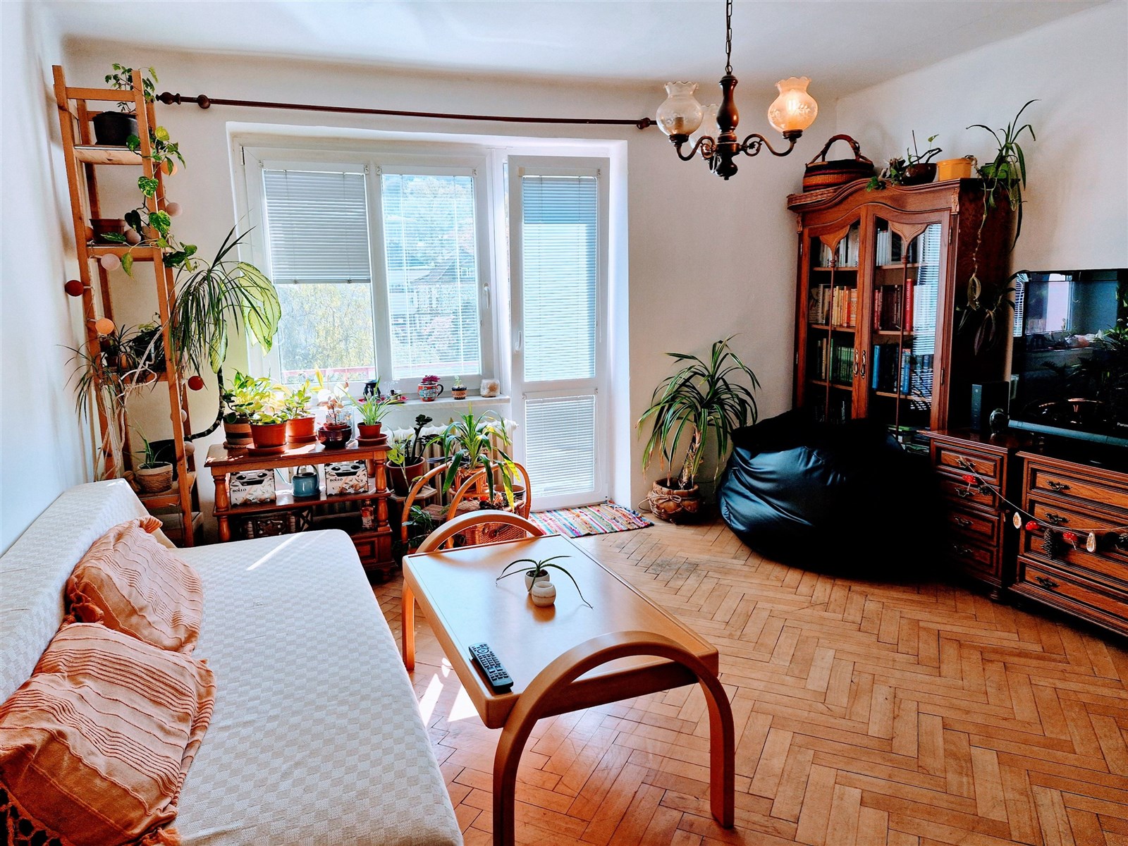 Světlý obývací pokoj s parketovou podlahou, velkými okny a rustikálním nábytkem, který poskytuje útulný a příjemný prostor pro relaxaci.