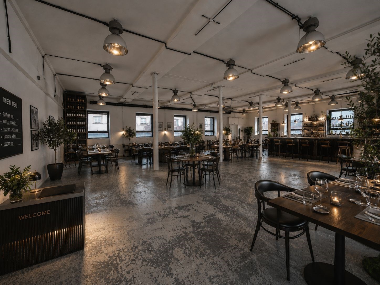 Prostorná restaurace s betonovou podlahou a industriálním designem, vybavená dřevěnými stoly a velkými okny, moderní osvětlení vytváří příjemnou atmosféru.