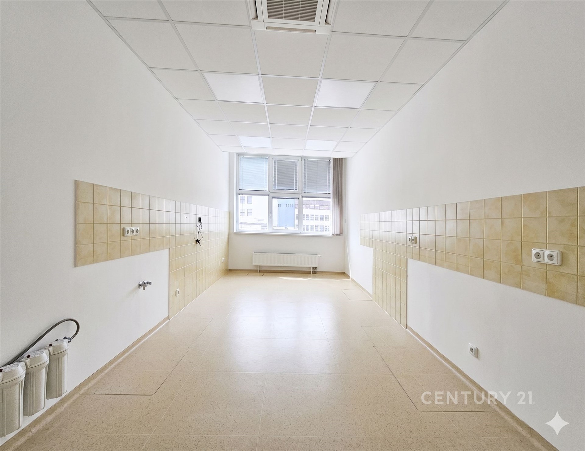 Prázdná kuchyně s dlažbou, připravená k rekonstrukci, disponuje velkými okny a bílými stěnami, ideální pro moderní úpravy.