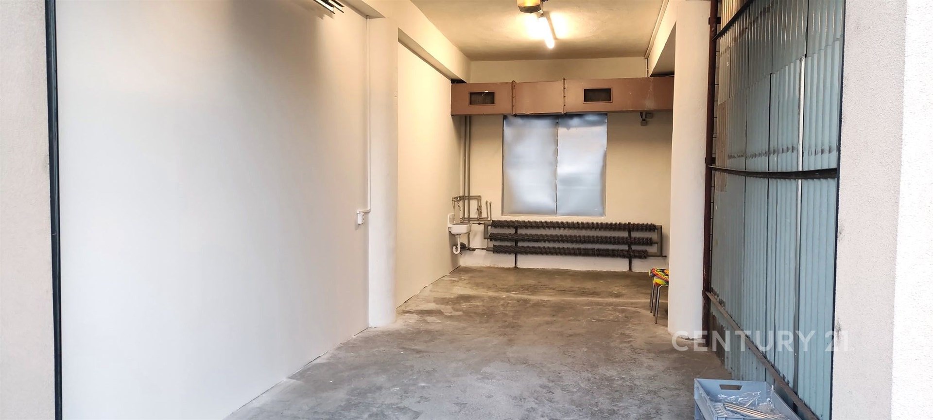 Úzká garáž se surovou betonovou podlahou, plechová vrata, bílými stěnami a stropním osvětlením, ideální pro parkování menšího auta.