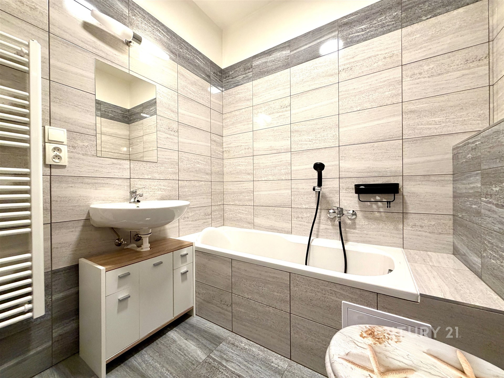 Moderní koupelna s dlažbou, vanou a umyvadlem, skříňka pod umyvadlem, koupelnový radiátor. Světlá rekonstrukce ve stylovém provedení.